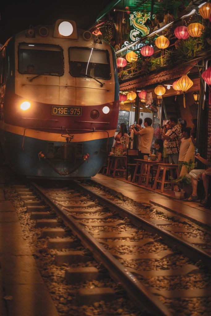 A train moves through a lively street cafe adorned with colorful lanterns at night.