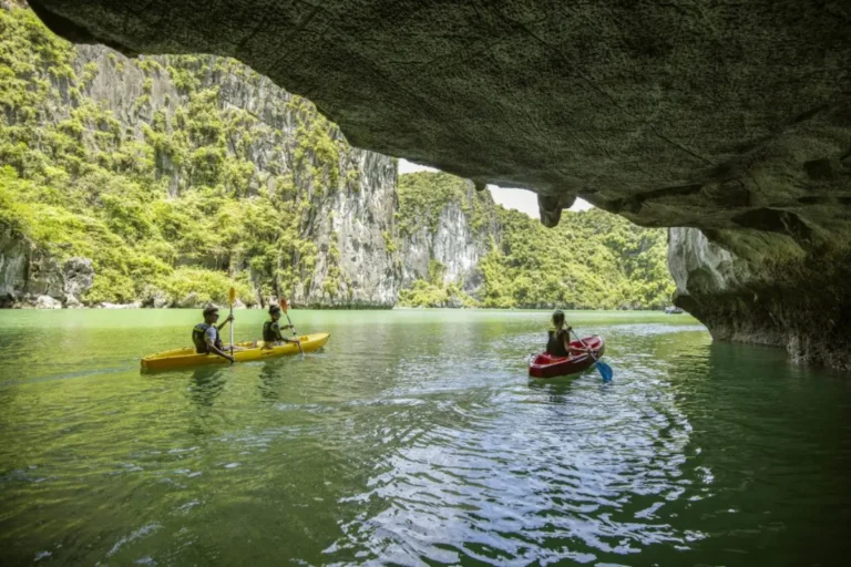 About Us 9 Travelers paddling kayaks out of a sea cave into a tranquil emerald lagoon in Halong Bay.