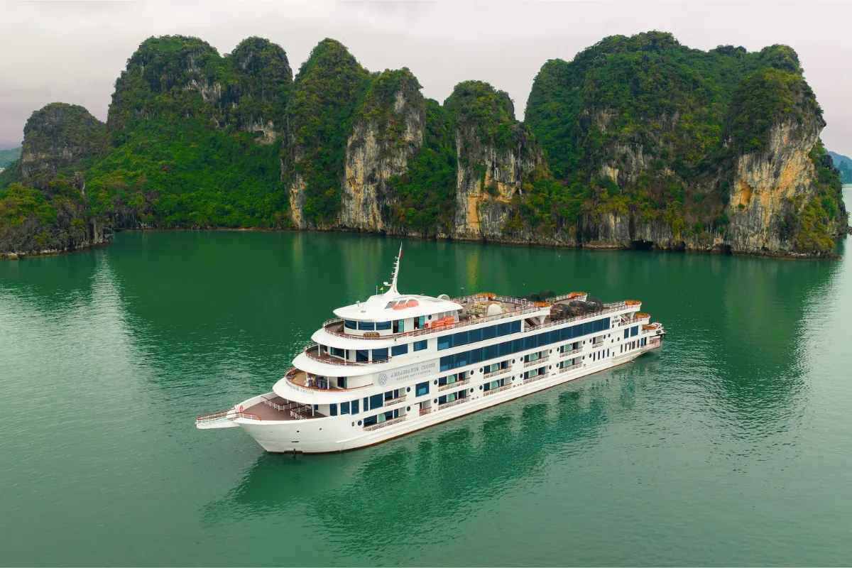 Ambassador Cruise luxury day tour ship cruising through emerald green waters past towering Halong Bay karsts.