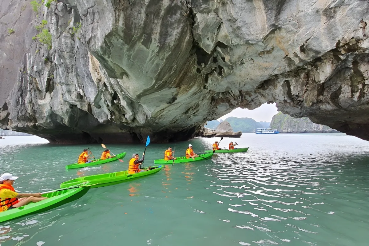 Kayakers paddling in a line through a narrow rock archway toward a hidden lagoon in Halong Bay