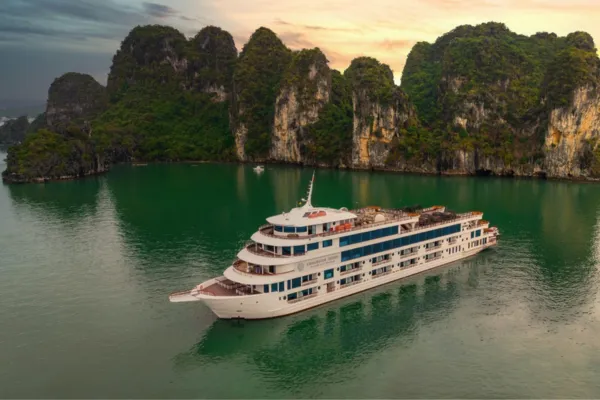 A multi-deck white cruise ship sailing past limestone karst islands in Halong Bay at sunset