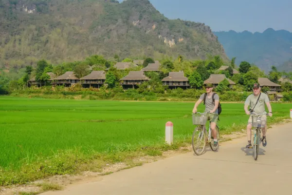 Two travelers cycling along a path beside green rice paddies and stilt houses in Mai Chau