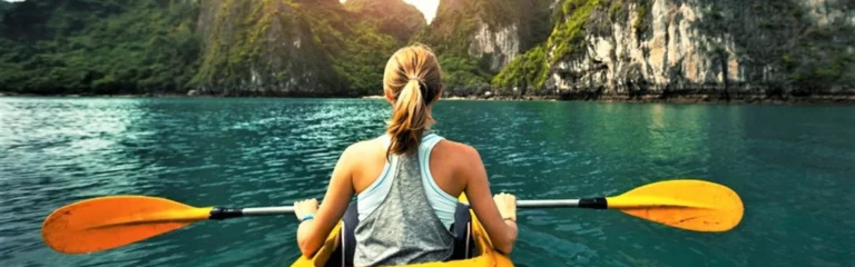 A woman kayaking toward towering limestone cliffs in the turquoise water of Lan Ha Bay