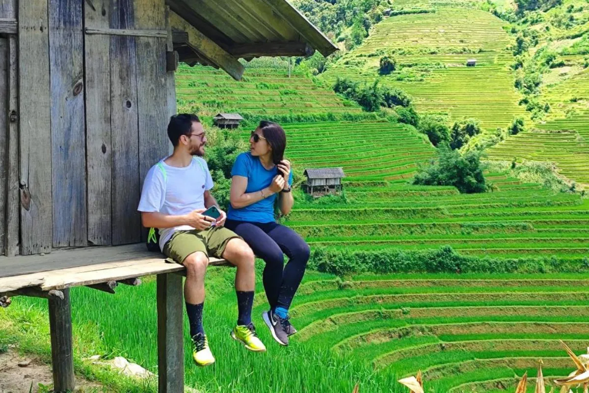 A couple sitting on a wooden hut platform overlooking layered green rice terraces in Mu Cang Chai