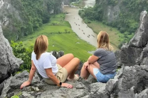 Two travelers sitting on the rocky summit of Mua Cave Viewpoint overlooking a winding river in Ninh Binh