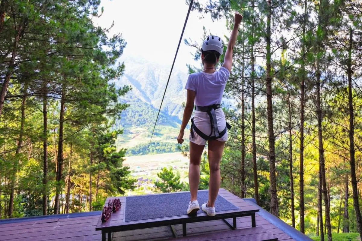 A woman on a zipline platform overlooking pine forest and mountains near Mu Cang Chai, arm raised before launching
