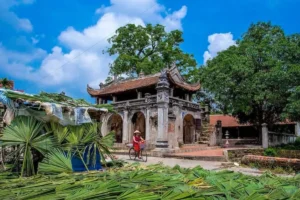 Woman cycling past Chuong Village's ancient stone gate, a Hanoi must-visit place for conical hats